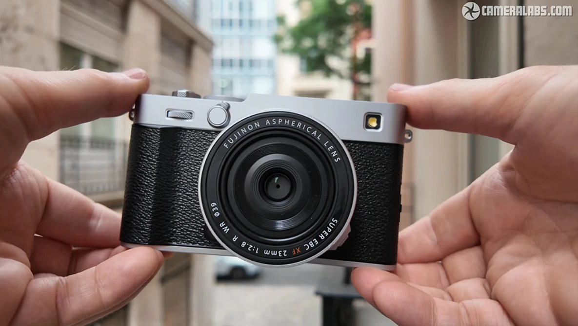 Hands holding a silver and black rangefinder camera with fixed lens against urban background.