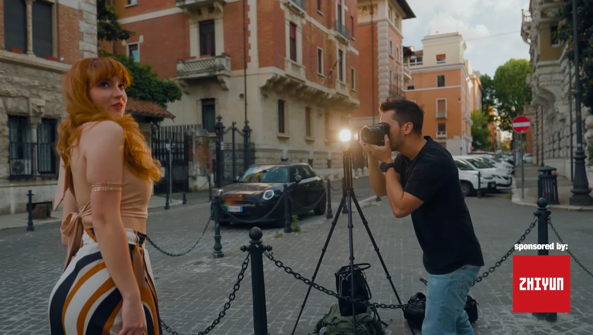 Photographer on tripod shooting a female model posing on a European cobblestone street with historic brick buildings.