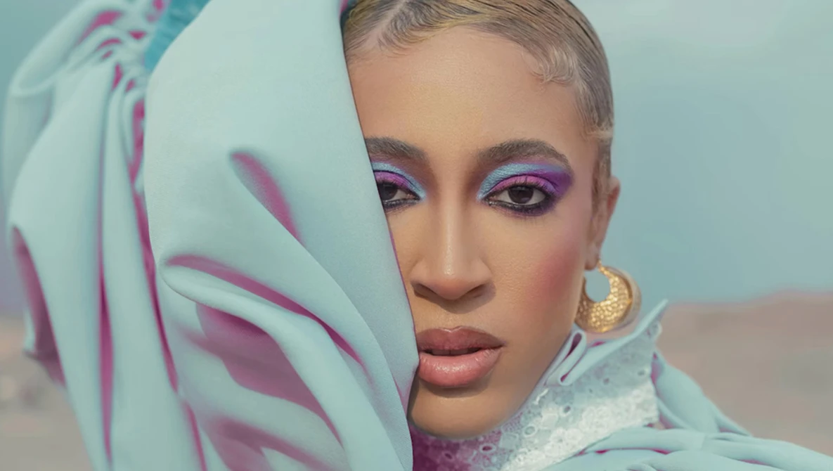 Woman with shaved head wearing purple eyeshadow and draped mint green fabric with magenta stripes.