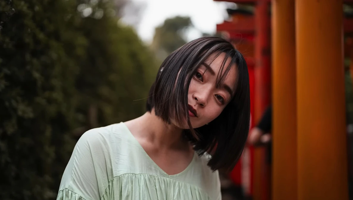 Young woman with short dark hair wearing a light green top, tilting her head with a contemplative expression against a blurred background with red and orange architectural elements.