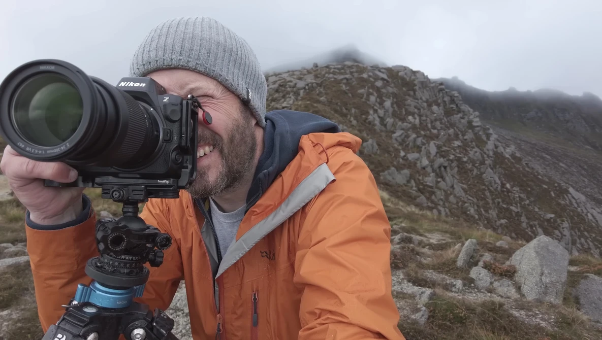 Photographer with Nikon camera on tripod looking through telephoto lens in mountainous terrain.