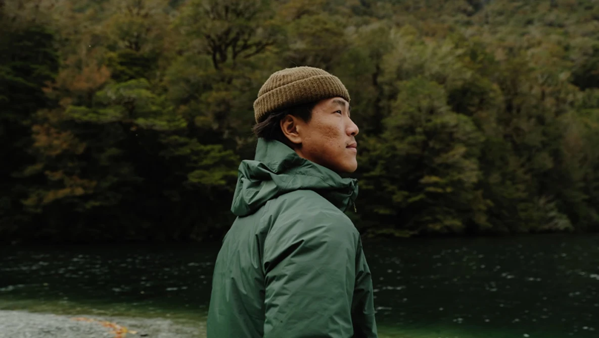 Person in green jacket and tan beanie looking toward a forested shoreline beside still water.