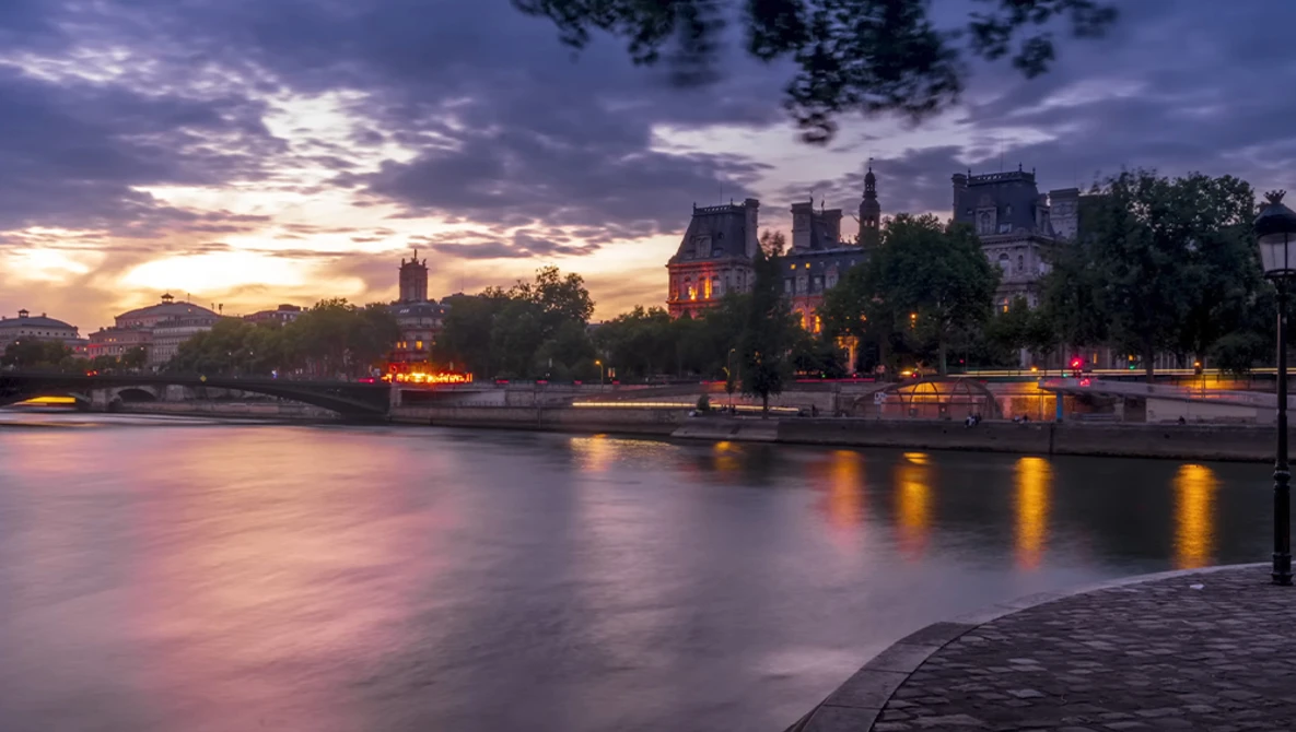 Illuminated riverside buildings at twilight reflected in still water under a dramatic purple and gold sky.