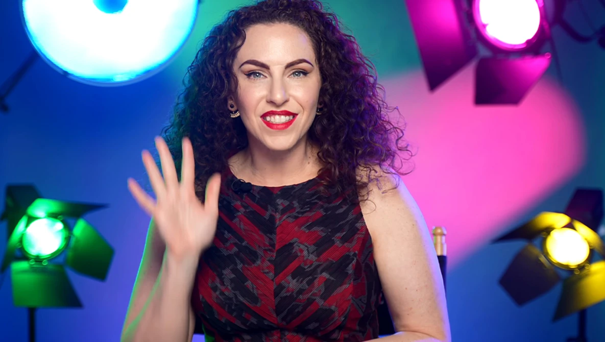 Woman with curly dark hair waving at camera against colorful studio lighting setup.