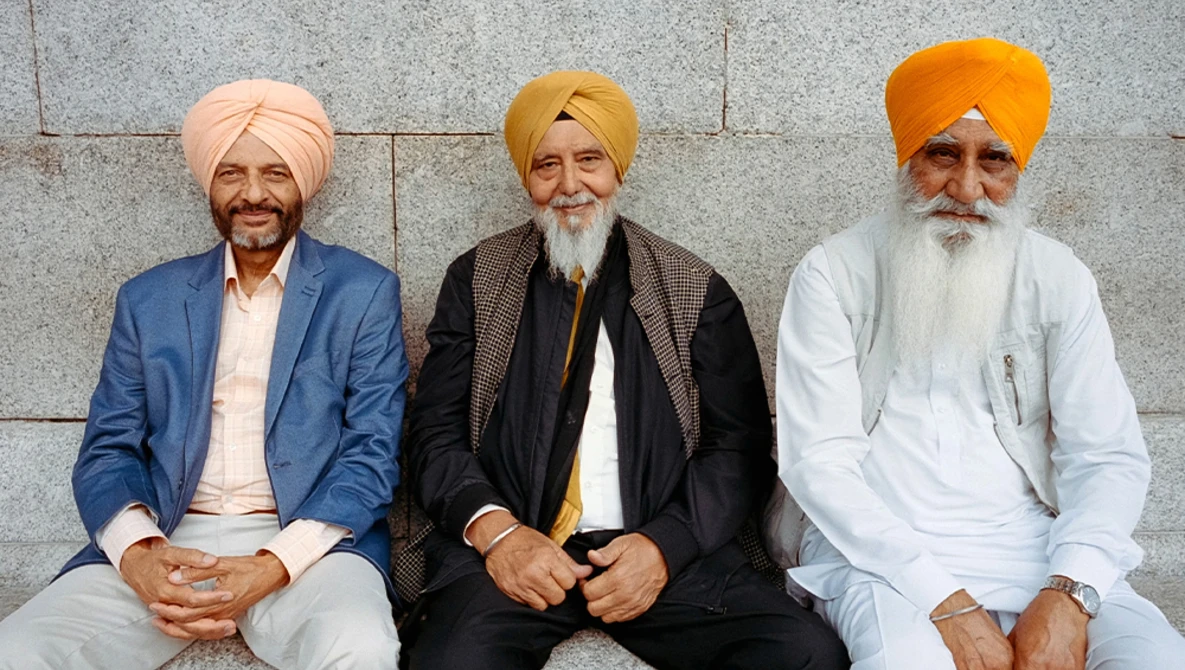 Three men with turbans seated against a stone wall, wearing formal jackets and traditional head coverings.