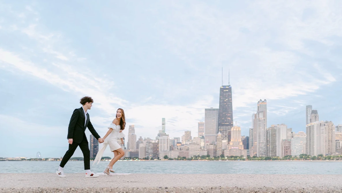 Couple in formal attire playfully running on a beach with a city skyline in the background.