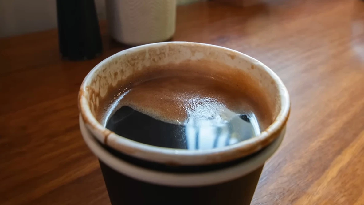 Iced black coffee in a ceramic cup on a wooden surface with a glass of milk visible in the background.