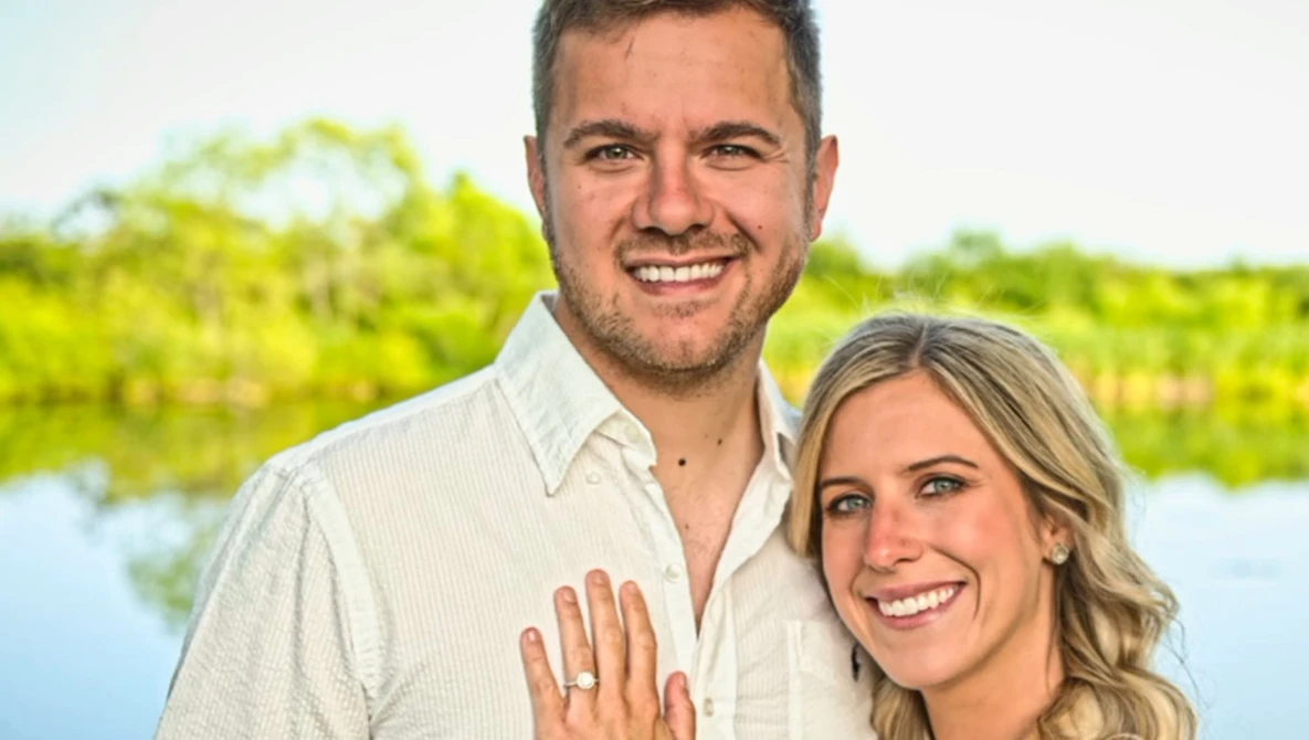 Smiling couple posing outdoors with water and green foliage in soft-focus background.