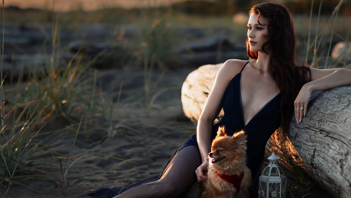 Woman in dark swimsuit seated on driftwood at beach during golden hour, holding small dog.