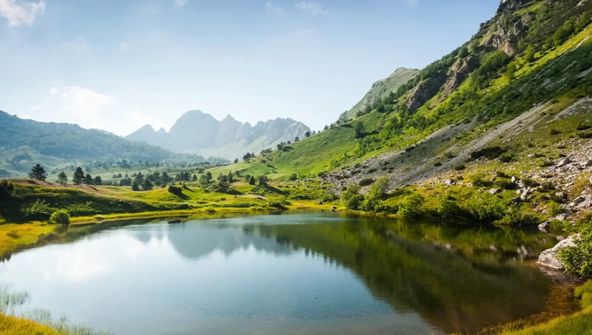 Alpine lake surrounded by green meadows and steep rocky mountains under clear sky.
