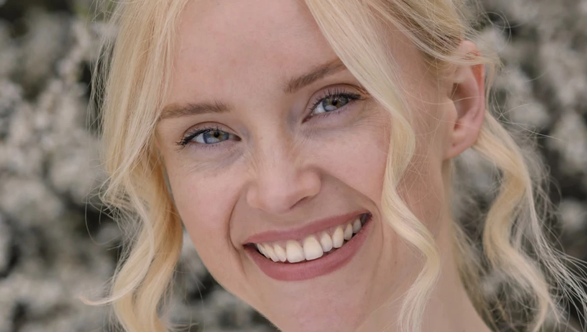 Blonde woman with wavy hair smiling at camera against textured gray background.