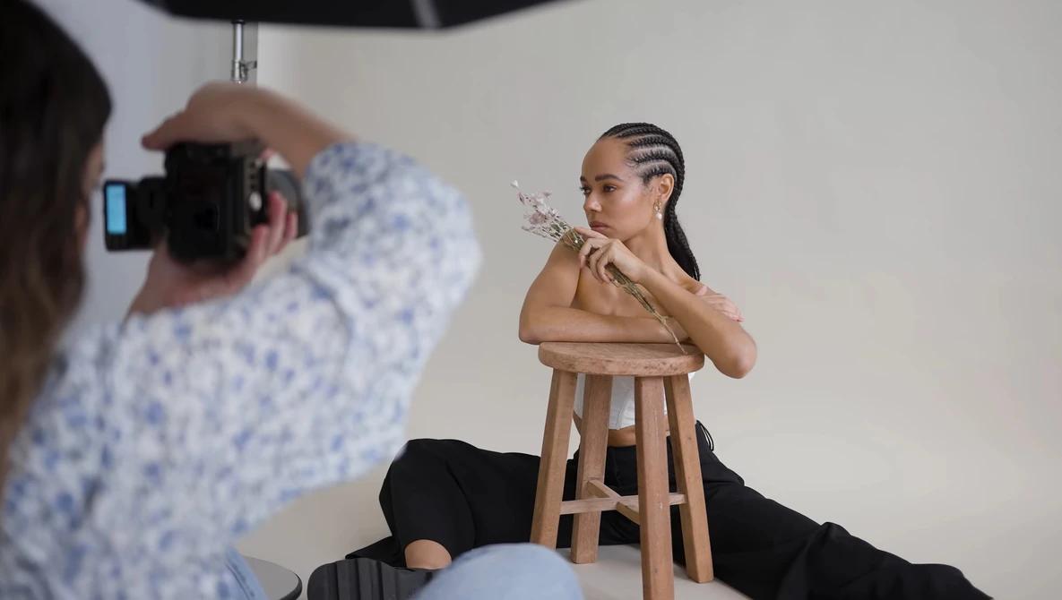Photographer shooting a portrait of a woman sitting on a wooden stool in a studio setting.