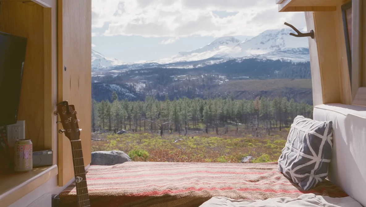 Bedroom window framing snow-capped mountain peak and forested valley landscape.