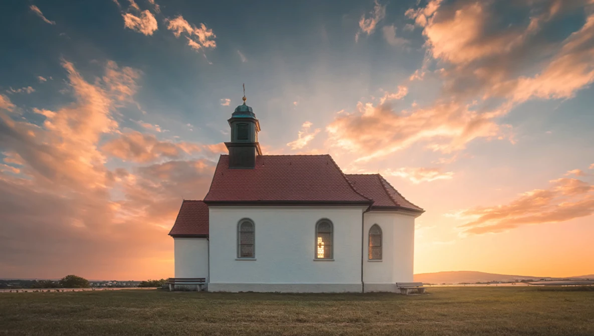 White rural church with red roof and steeple standing alone in an open field at sunset.