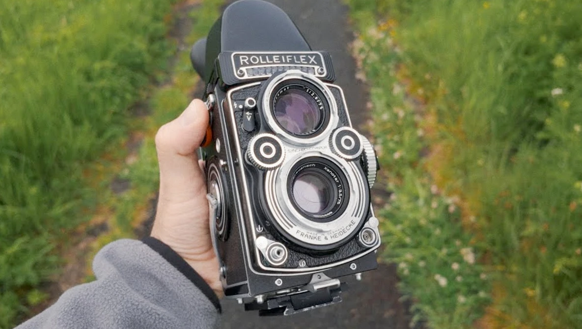 Rolleiflex twin-lens reflex camera held in hand against a blurred green field background.