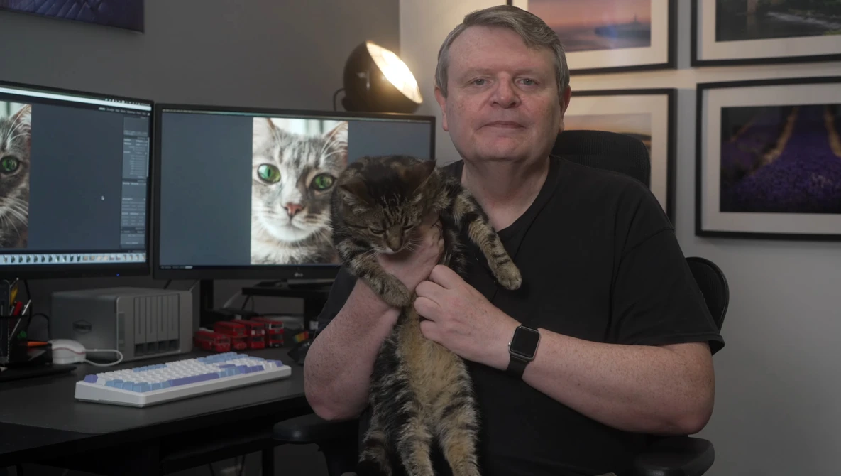 Man holding a tabby kitten in a photography studio workspace with dual monitors displaying cat images.