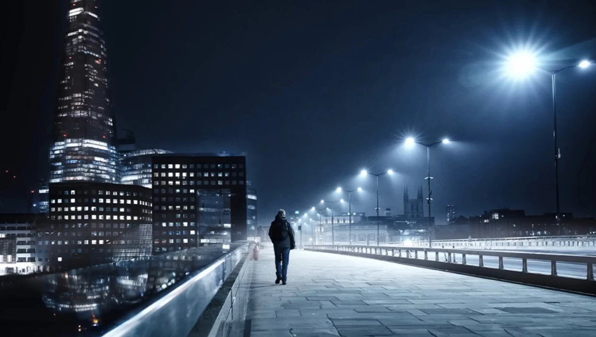 Solitary figure walking across an illuminated bridge at night with a modern city skyline in the background.