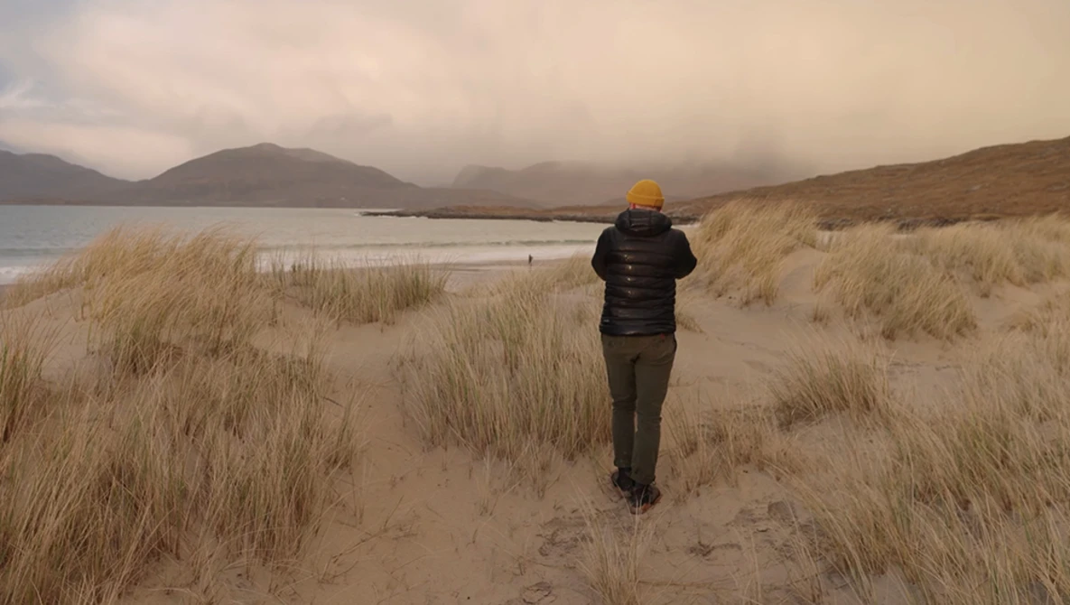 Runner in yellow hat jogging through sandy beach with dune grass toward misty mountains and water.