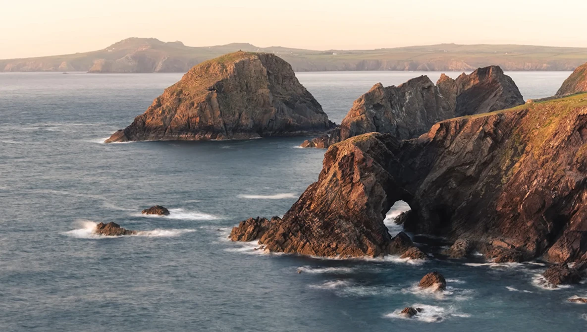 Rocky coastal cliffs with a natural arch opening to the sea, bathed in golden hour light.
