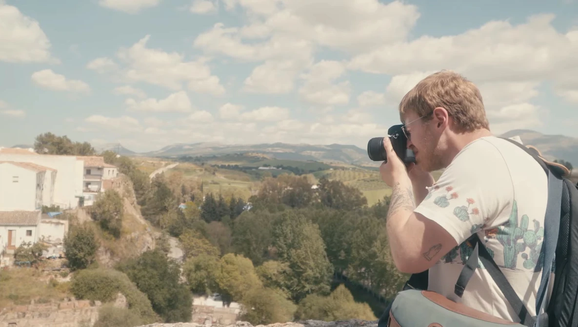 Photographer holding camera to face, composing a shot of rolling hills and forested landscape.