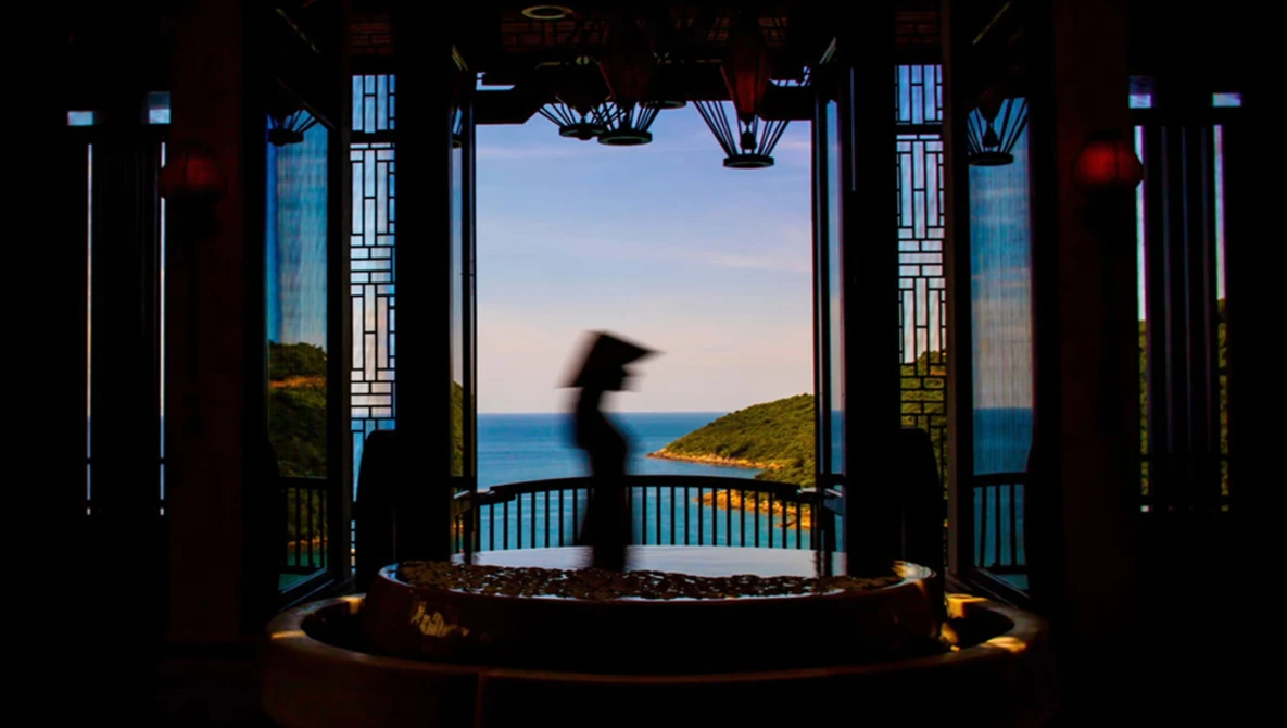 A blurred figure moves through an ornate pavilion overlooking a coastal landscape at twilight.