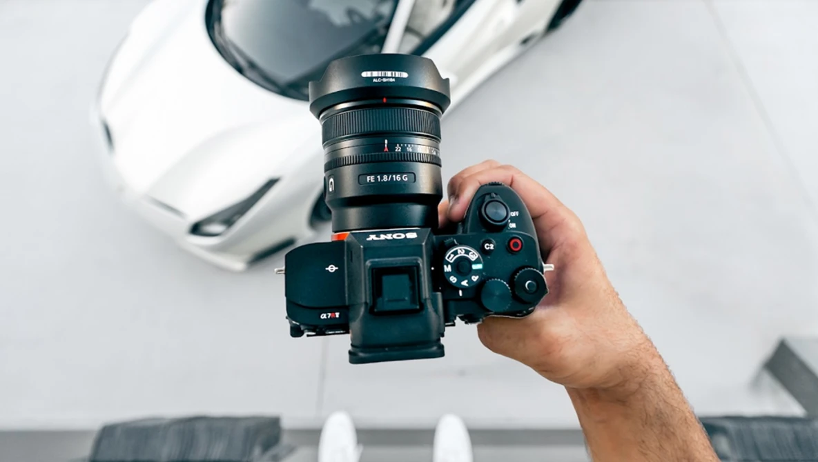 Photographer holding a mirrorless camera with mounted lens and external flash against a white backdrop.