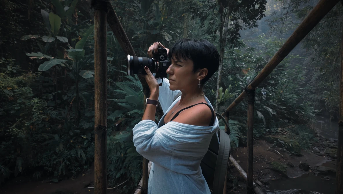 Woman in light blue off-shoulder dress holding vintage film camera in lush forest setting.