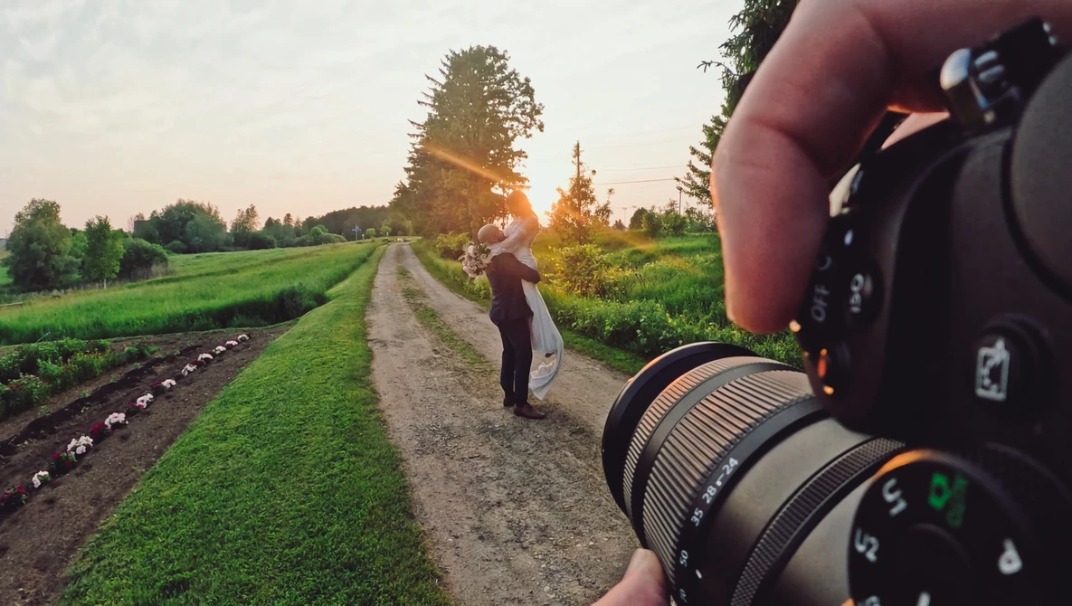 Photographer holding camera with telephoto lens, photographing person standing on rural dirt road at golden hour.