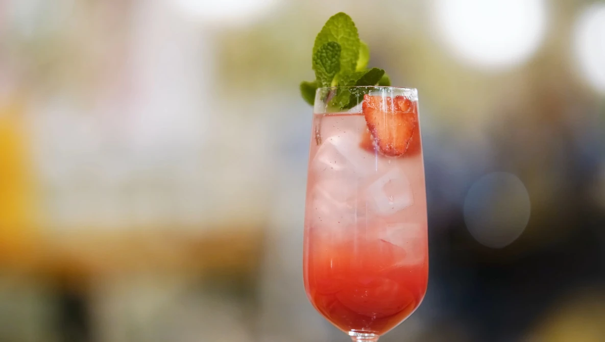 Red cocktail with ice, mint garnish, and strawberry slice in tall glass.