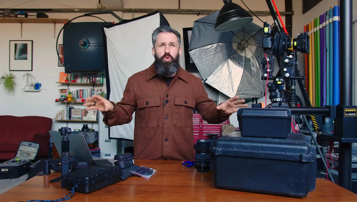 Bearded man in burgundy shirt standing at wooden desk surrounded by photography lighting equipment and cases.