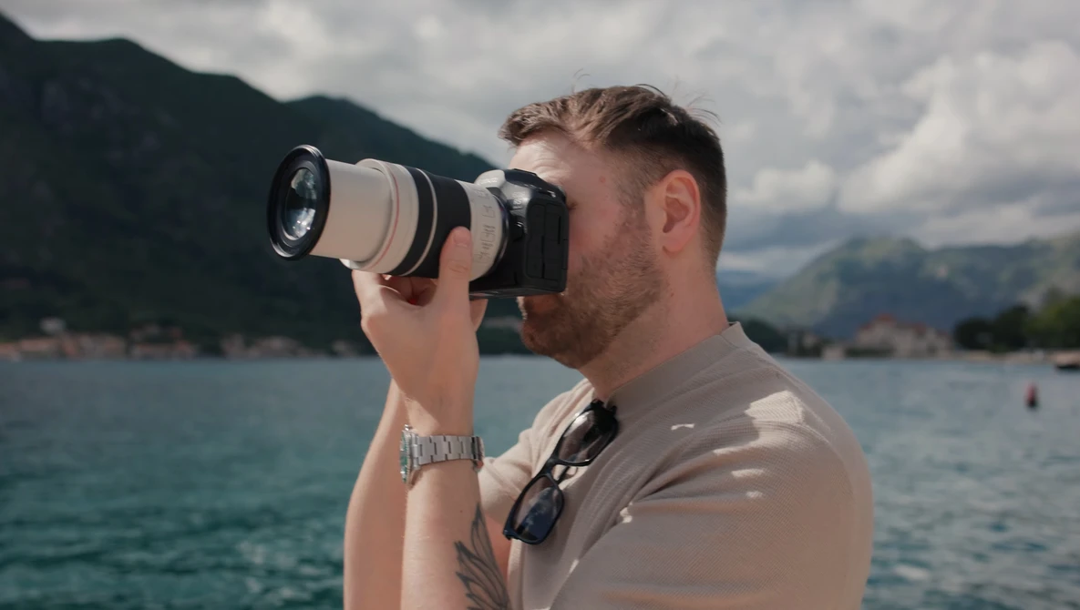 Photographer with telephoto lens photographing across a lake toward mountains on a partly cloudy day.