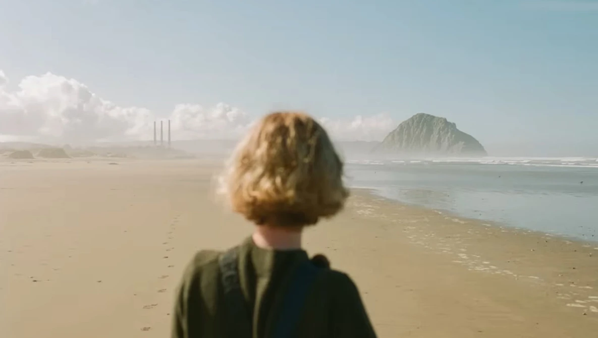 Person with blonde hair viewed from behind standing on a wide sandy beach, gazing toward a distant rocky island and industrial structures.