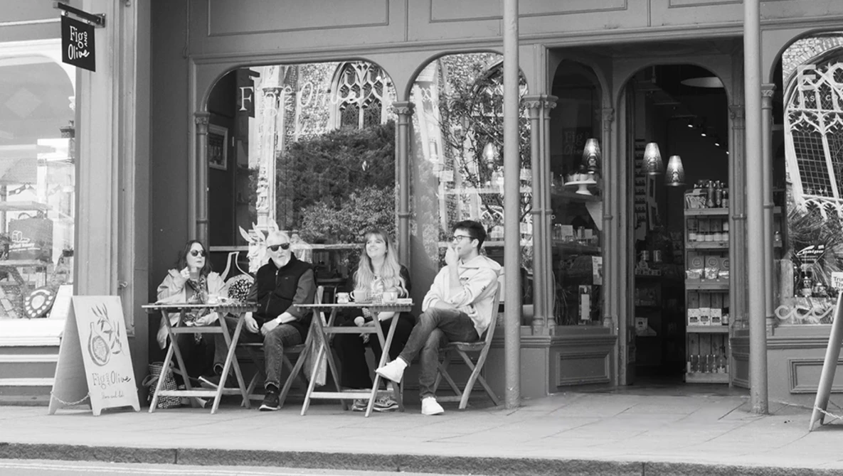 Four people seated outside a storefront with large windows, conversing in black and white.
