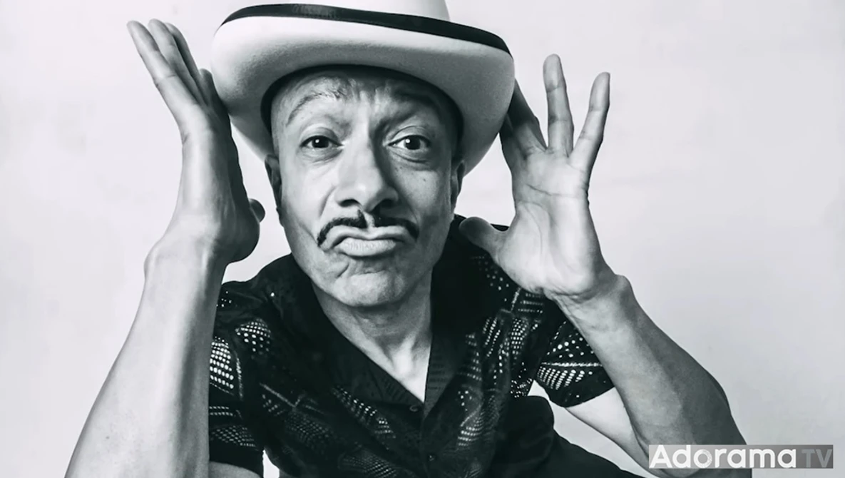 Man in cowboy hat and patterned shirt with hands raised to sides of head, black and white portrait.