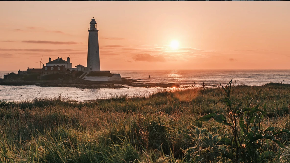 Lighthouse silhouetted against golden sunset sky with keeper's house and coastal grassland in foreground.