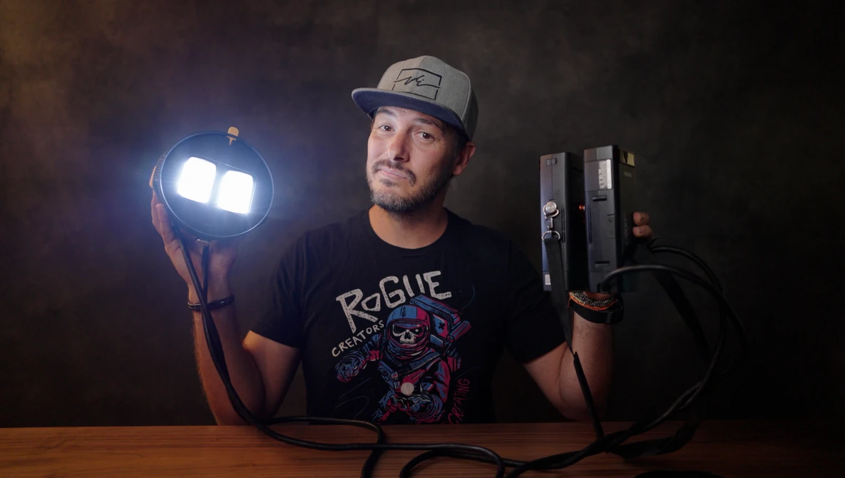 Photographer holding a portable LED light panel in one hand and lighting equipment in the other against a dark background.