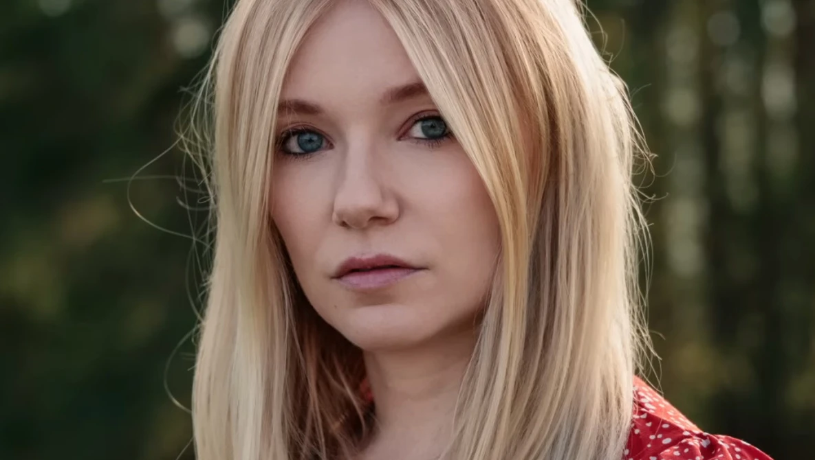 Portrait of a woman with platinum blonde hair and blue eyes against a blurred forest background.
