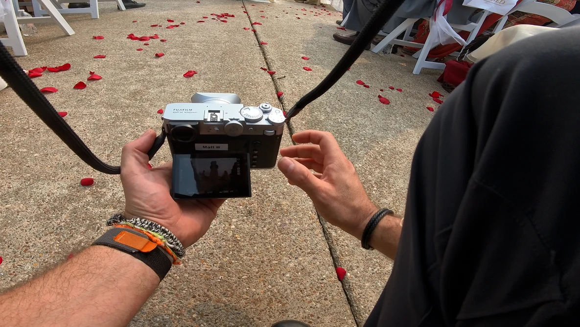 Hands holding a vintage rangefinder camera with a cable release attached, outdoors at an event with red petals on the ground.