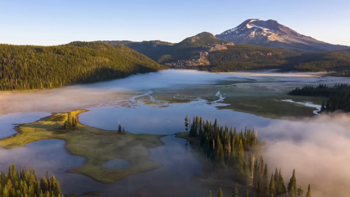 Snow-capped volcanic peak rising above forested valleys with layered lakes and morning mist.