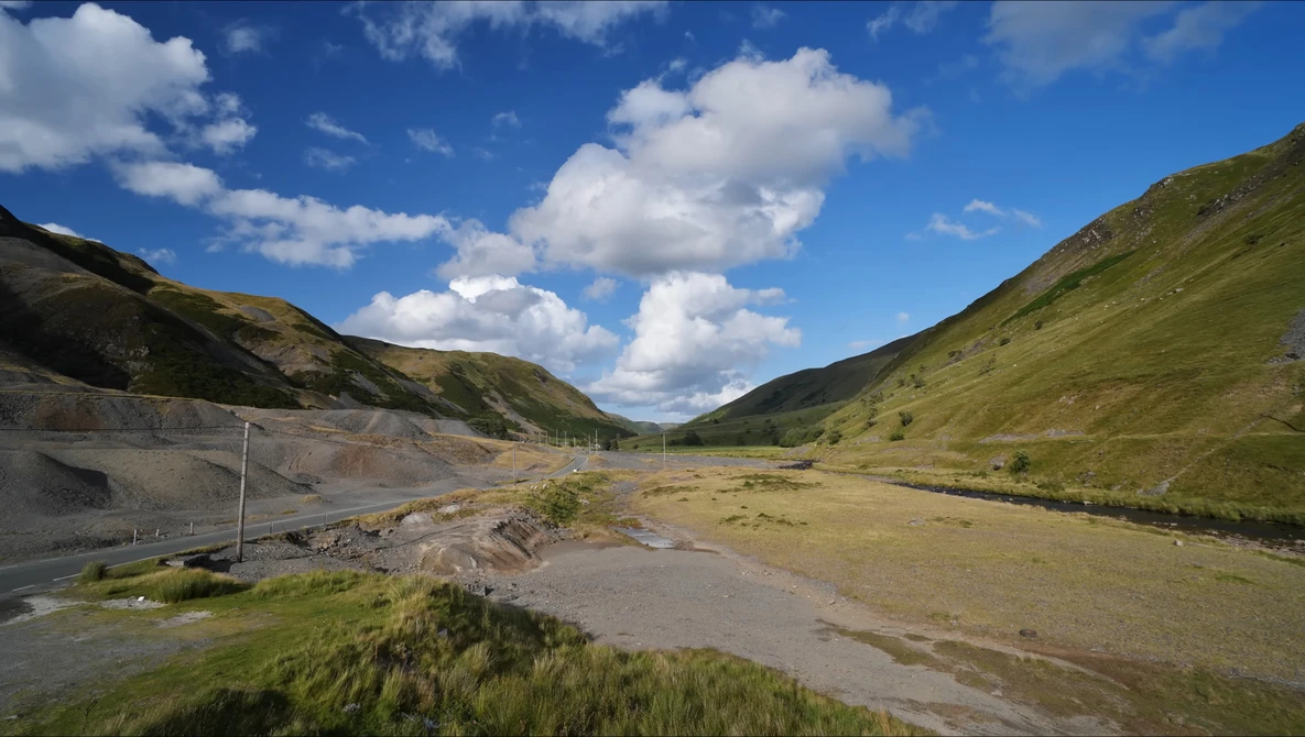 Alpine valley with braided river, grassy slopes, and dramatic sky with white clouds.