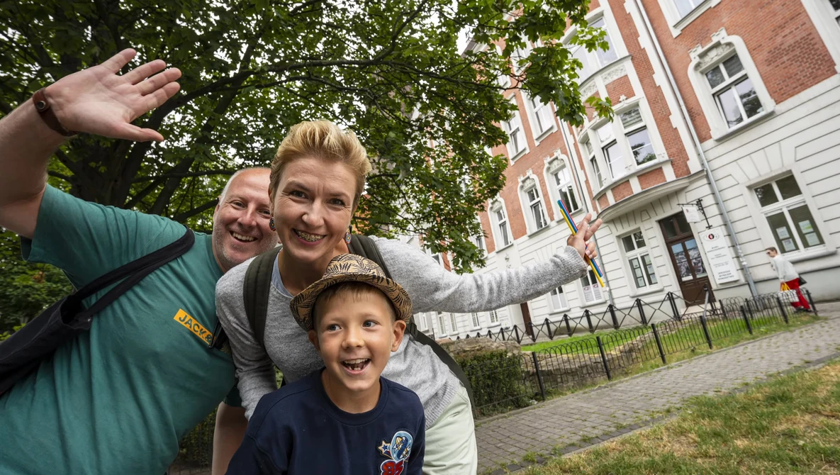 Three people posing together outdoors with arms raised, smiling at the camera in front of a brick building.