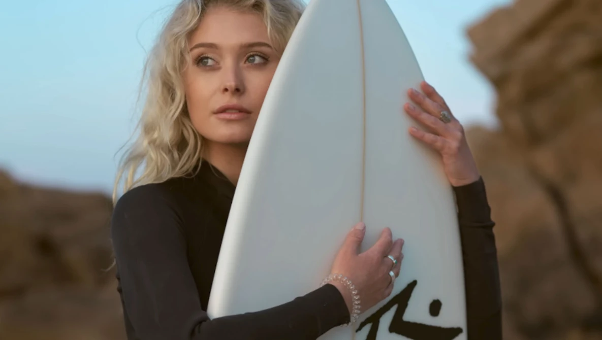 Blonde woman in black wetsuit holding white surfboard at rocky coastline.