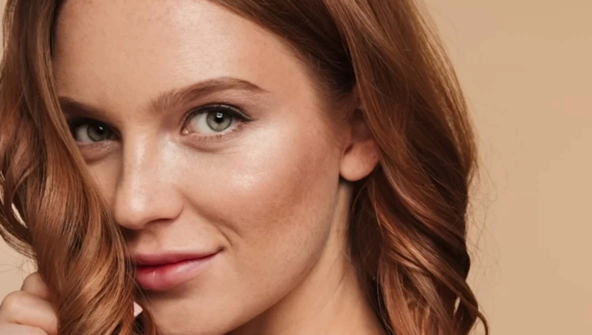 Woman with wavy auburn hair and green eyes against a warm beige background.