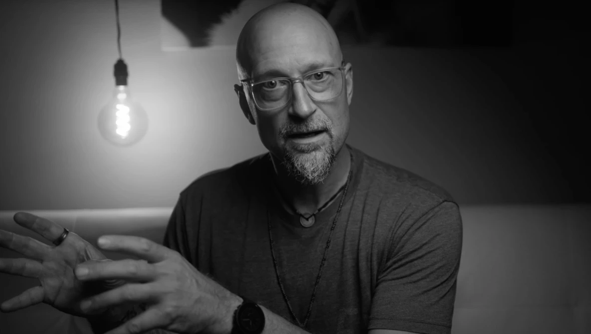 Man with glasses wearing a dark t-shirt gestures while speaking, lit by a hanging incandescent bulb against a dark background.