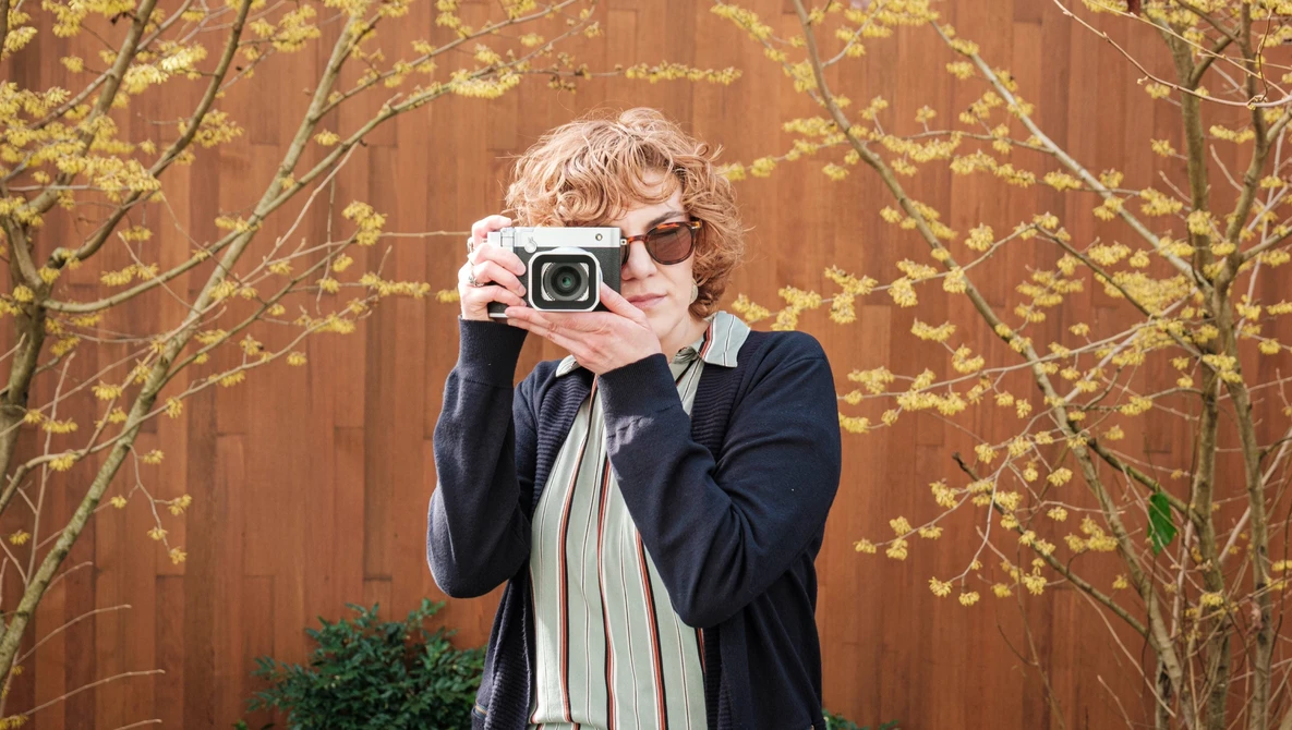 Photographer with gray curled hair and sunglasses holding a vintage film camera to her face.