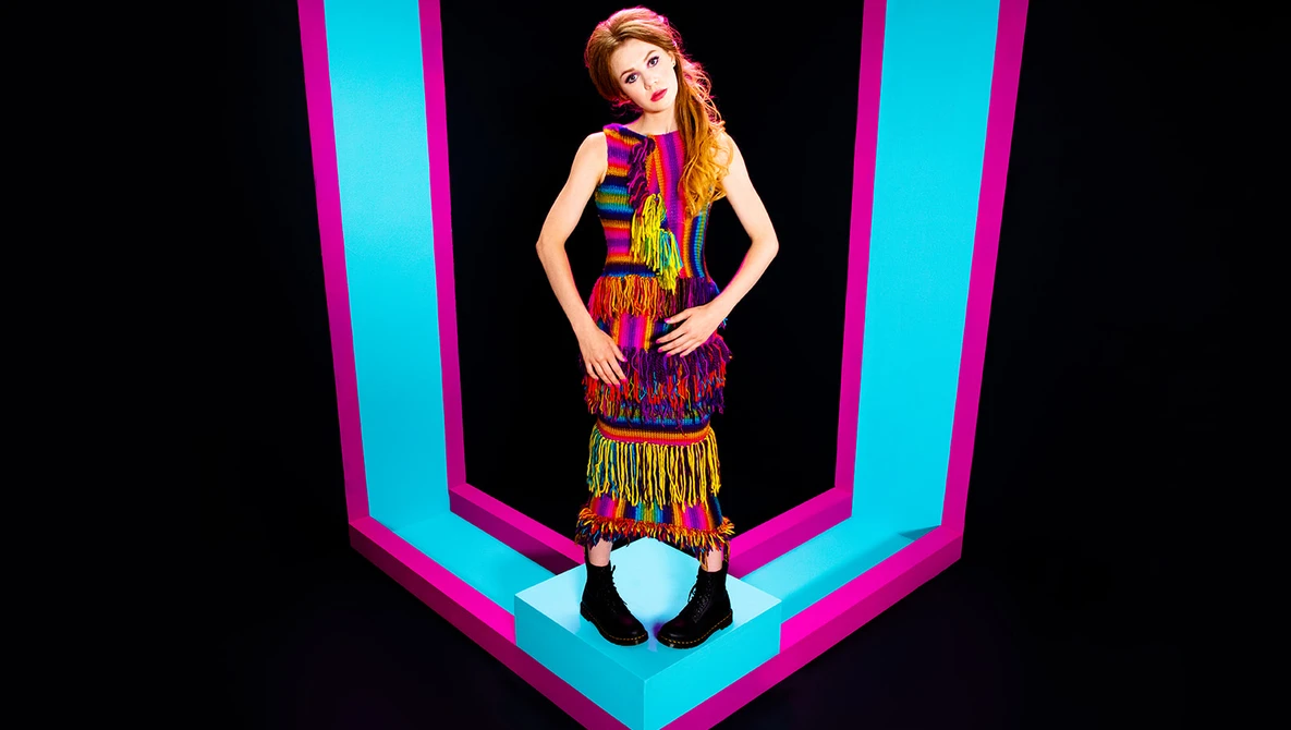 Woman in colorful fringed outfit posed against bright cyan and magenta geometric neon lines.