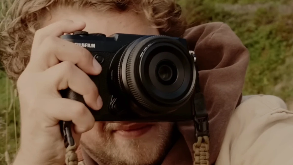 Photographer holding a Fujifilm mirrorless camera up to his face to compose a shot outdoors.
