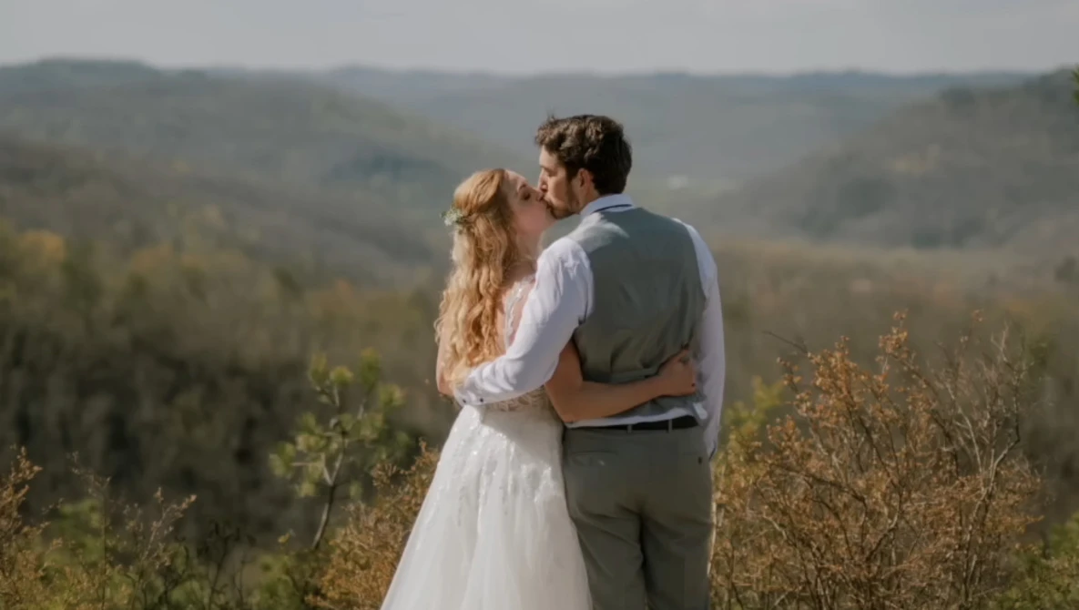 Couple in wedding attire kissing on a hillside overlooking forested valley.