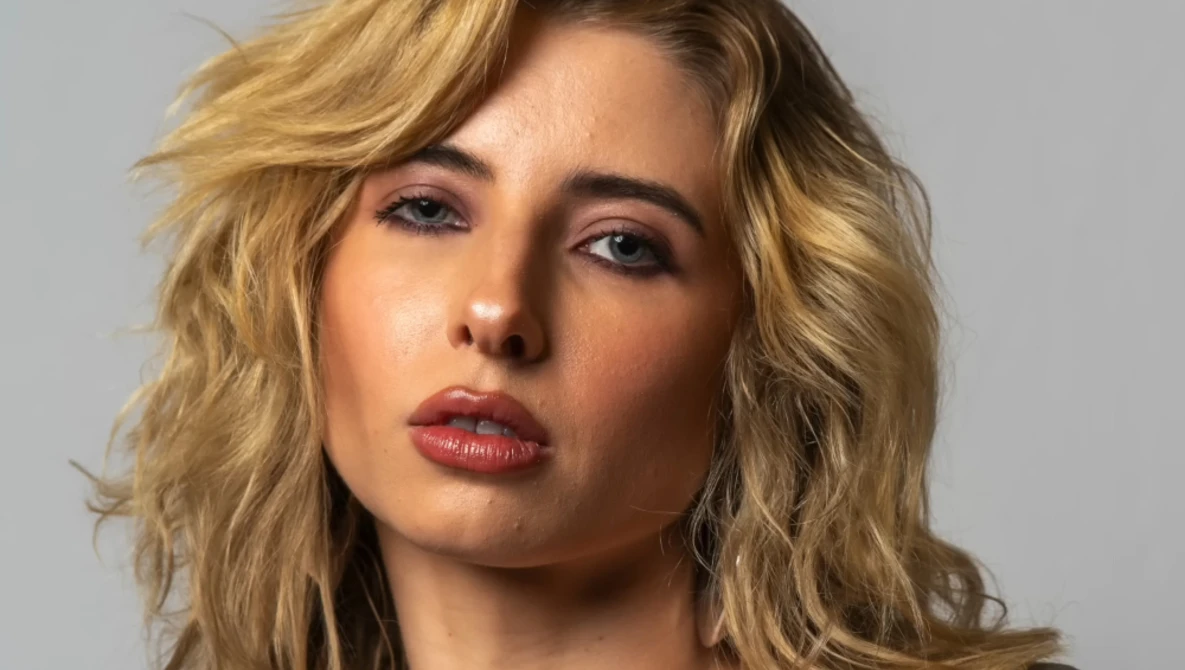 Portrait of a woman with shoulder-length wavy blonde hair against a neutral gray background.