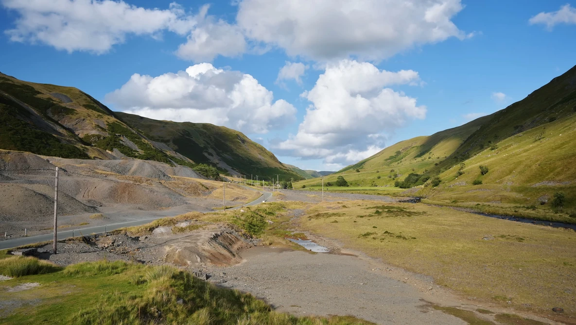 Mountain valley with grassy slopes, exposed earth, and a small stream under blue sky with white clouds.